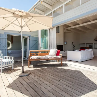 Pebbles home with beige clapboarding and blue double wooden doors that open a bedroom suite onto the expansive wooden deck with an outdoor wooden bench and an armchair shaded by a white umbrella and large glass stack-back doors that open the lounge onto the deck.