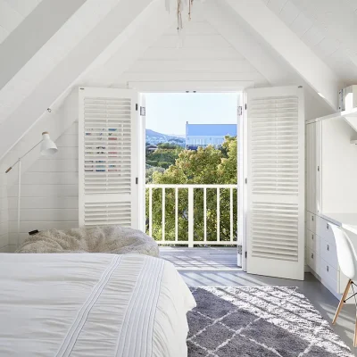 Loft bedroom with a built in cupboard and desk, soft grey epoxy flooring, a spacious bed with crisp white linen, a large bean bag and double doors that open onto a wooden deck with views of greenery. Noordhoek Beach House.