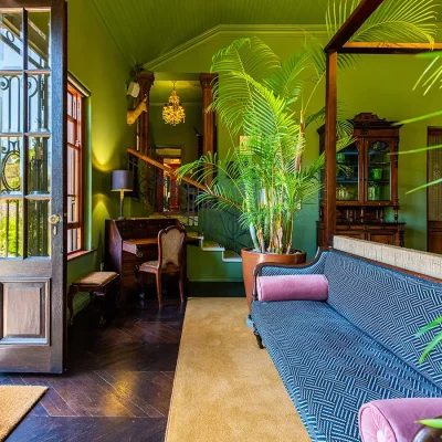 Bright green bedroom with four poster bed, sofa and palm trees looking out wooden double doors. House Nouveau.