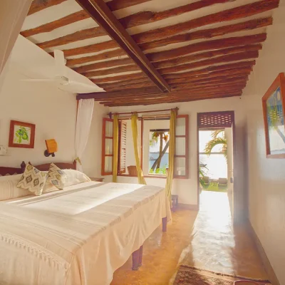 First floor bedroom suite opening onto an ocean view patio, with cream hues in the linen, walls and curtains that compliment the wooden bed frames and ceiling. Peponi Hotel.