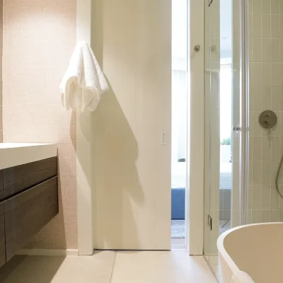 Ebb Tide en-suite bathroom with modern bath tub, an enclosed- glass shower, a long sleek wooden bathroom cabinet and white counter tops and mounted oval mirrors on a cream textured wall.