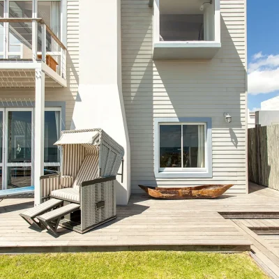 Beige cladded beach house with blue window frames and a wrap around deck with a large shaded reclining armchair with striped cushioning. Pebbles.