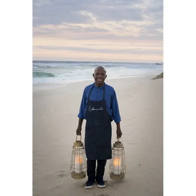 A staff member at Lekkerwater beach lodge standing and smiling . on the beach with two lanterns in hand.