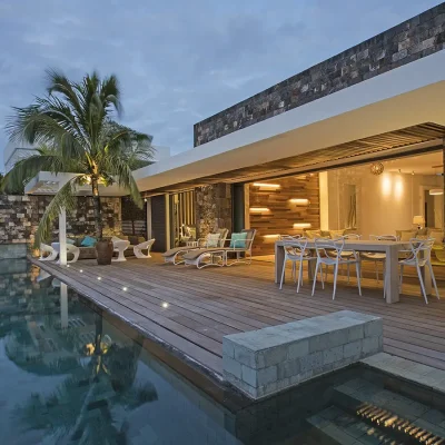 Evening hues of a warmly lit up villa opening onto a secluded deck with outdoor dining furniture and a large pool.