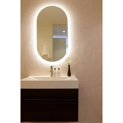 Ebb Tide bathroom with lit-up oval mirror mounted against a cream, textured wall with a single ceramic sink with a vase of white flowers and attached to a wooden cabinet.