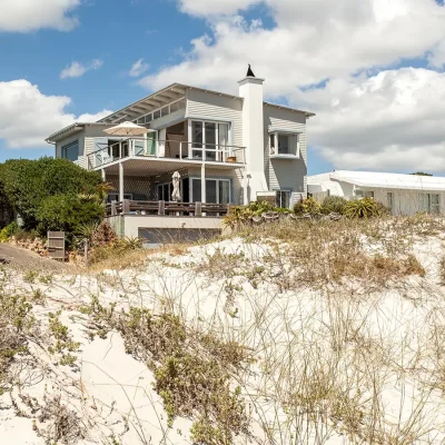 Pebbles home with beige clapboarding, a wrap around deck on the first level and a balcony on the second level, perched behind sand dunes.