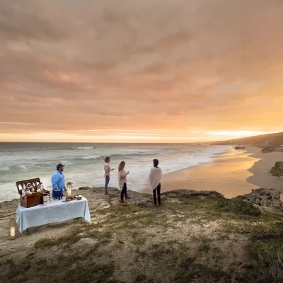 Sundowners on the coastal plateau with couples standing with drinks in hand watching the sky turn orange across the stretch of De hoop nature reserve coastline.