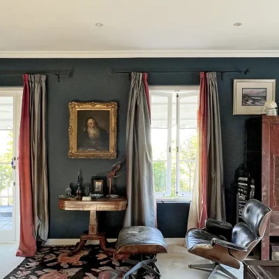 Bedroom with navy walls decorated in art, large windows and glass doors with rugged silver and red curtains, a black and red carpet and an elongated leather armchair. Villa Toscana.