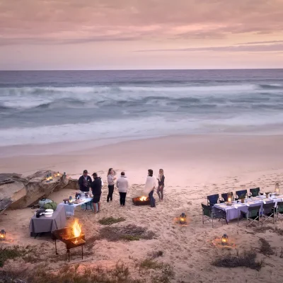 A group of people on the beach surrounded by fire pits, and a long dining table overlooking the sunset on the ocean. Lekkerwater Beach Lodge.