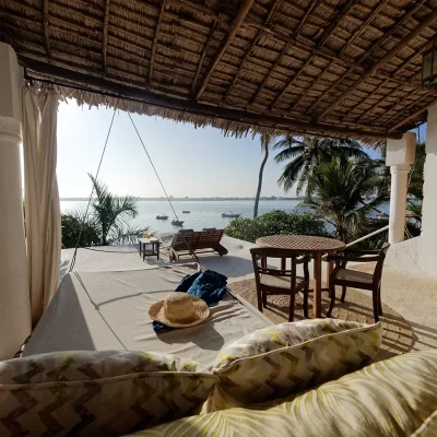 Outdoor swinging bed on a bedroom patio with a wooden table and chairs shaded by a thatched roof and looking onto the ocean.