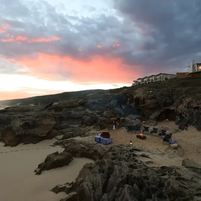 A moody evening on the beach warmed by hints of orange from the setting sun and the large fire pit surrounded by rock formations and Lekkerwater beach lodge on the horizon.