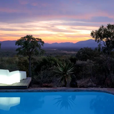 Sunset from the lit up swimming pool at Manor House overlooking the terrain and sky with shades of pink, purple and orange.