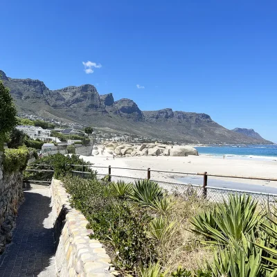 Paved side walk adjacent to Glen beach with white soft sand and blue ocean water with the Twelve Apostle mountain range in the distance.